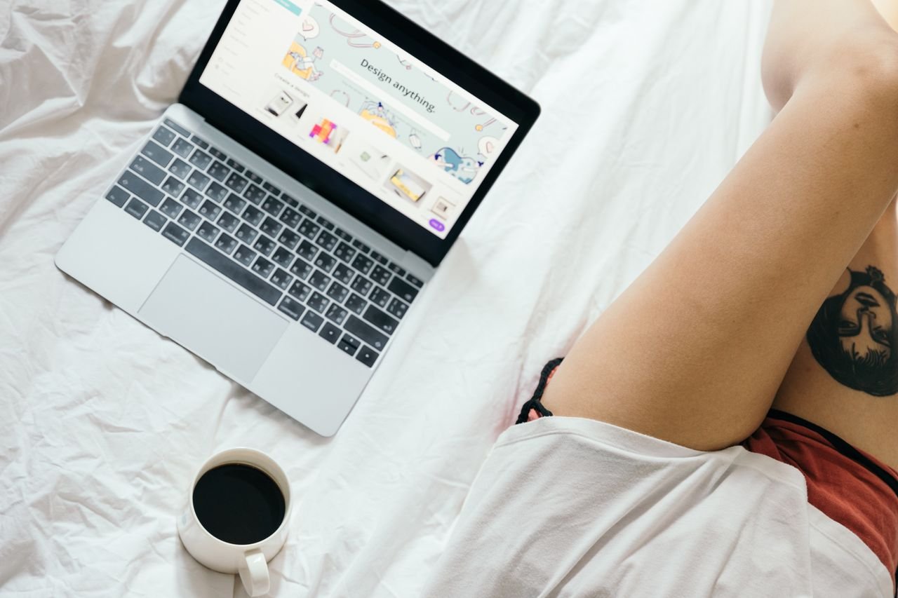 Home From above of unrecognizable woman with tattoo on leg browsing netbook and drinking coffee while resting on bed in cozy bedroom
